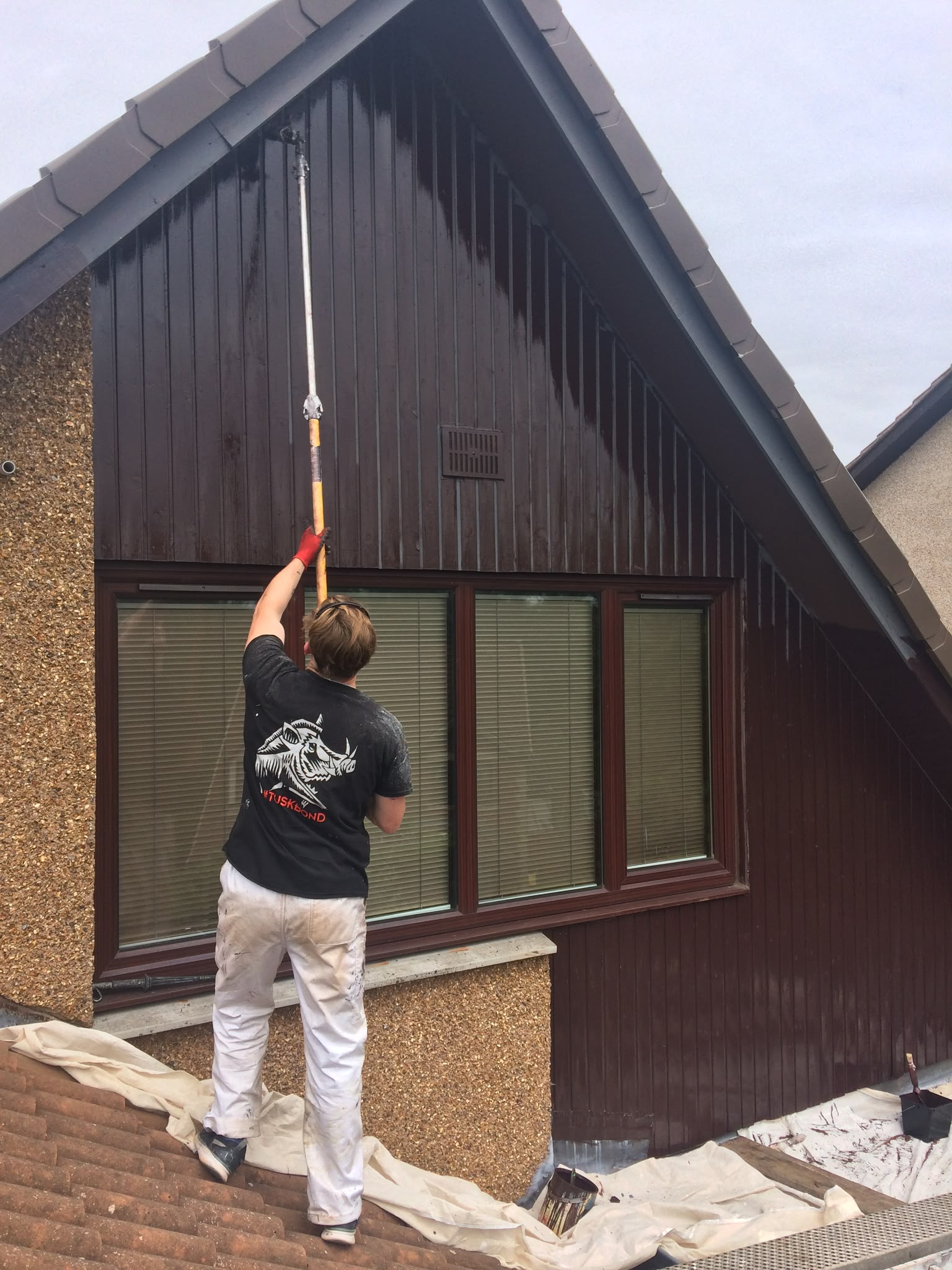 Painter applying paint to brown timber cladding on a house gable.